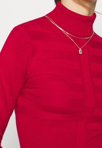 Pull à col roulé rouge avec un motif texturé à rayures horizontales, col côtelé, et deux colliers dorés superposés.