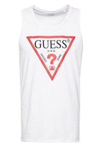 Débardeur en coton blanc arborant un grand logo triangulaire rouge avec le texte "GUESS?" et "U.S.A. WASHED JEANS" en noir. Texture lisse, design sans manches.