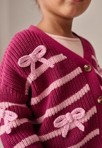 Fuchsiová a svetloružová pruhovaná cardigan so V-výstrihom, zdobená pletenými mašľami na pleciach a veľkými gombíkmi. Textúra je rebrovaná.