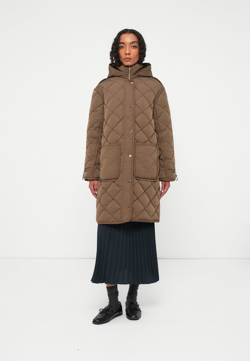 Casaco longo castanho acolchoado com capuz, apresentando dois bolsos frontais e botões de pressão, combinado com uma saia plissada escura e sapatos pretos.