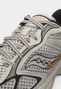 Κοντινό πλάνο γκρι και άσπρου αθλητικού παπουτσιού Saucony με υφασμάτινο πλέγμα, μαύρο και πορτοκαλί λογότυπο και λεπτομέρειες στα άσπρα και μαύρα κορδόνια.