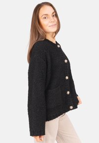 Schwarze, flauschige Strickjacke mit rundem Ausschnitt, vorderen Taschen und sechs metallischen Knöpfen. Der Stoff hat eine strukturierte, gemütliche Optik.