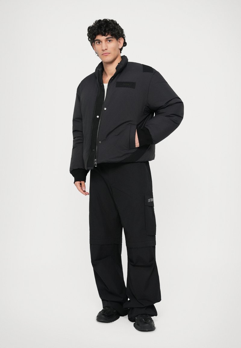 Schwarze Steppjacke mit hohem Kragen, Fronttaschen und schwarzen Akzenten. Kombination mit lockeren schwarzen Hosen und dunklem Schuhwerk.