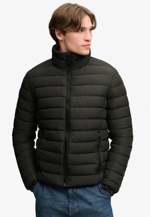Hombre que lleva una chaqueta acolchada negra con bolsillos con cremallera y vaqueros azules, mirando hacia la derecha con las manos en los bolsillos.