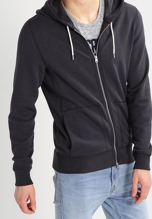 Mężczyzna w czarnej bluzie z kapturem zapinanej na zamek z białymi sznurkami i jasnoniebieskich dżinsach, stojący na tle jednolitego jasnego tła.