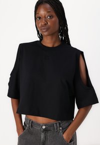 Camiseta negra cropped de algodón, con mangas cortas y detalles recortados, y logo en relieve en el frente. Combinada con pantalones grises.