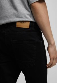 Vaqueros de mezclilla negra con un acabado texturizado. Presentan un parche de cuero marrón con la etiqueta "PULL&BEAR" en la parte trasera de la cintura. Diseño sencillo de bolsillos.