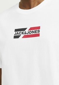 Hvid bomulds t-shirt med trykt logo i sort og røde accenter, korte ærmer og rund hals.