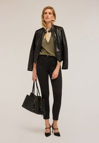 Mujer de pie con chaqueta de cuero negra sobre una camiseta verde brillante, pantalones skinny negros, tacones negros y llevando un gran bolso negro.
