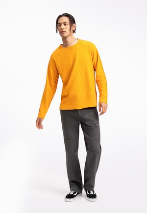 Felpa gialla a maniche lunghe con scollatura rotonda, abbinata a pantaloni cargo grigi con tasche laterali e vestibilità comoda. Inclusi sneakers nere.