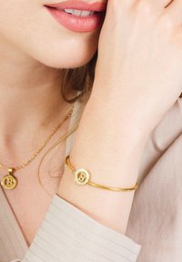 Gouden armband met een ronde charm met een verhoogde letter "B." Glad metaaltextuur, slank ontwerp, gedragen op de onderarm met een shirt in neutrale tinten.