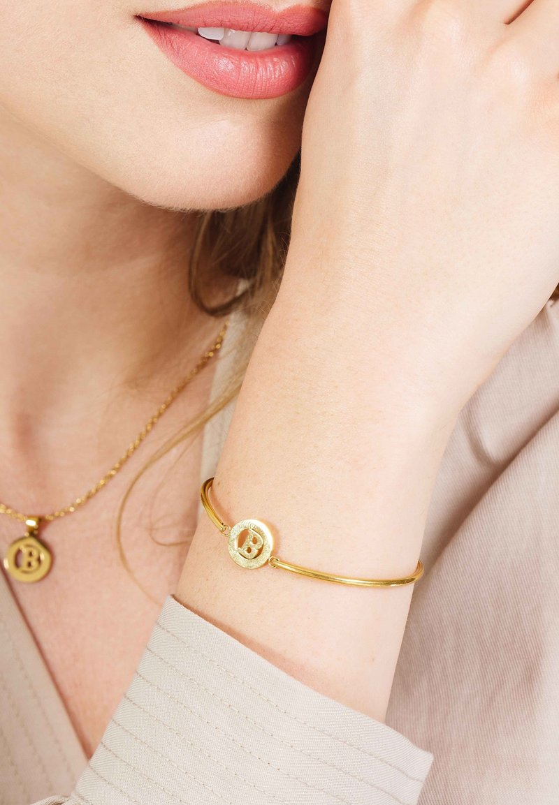 Gouden armband met een ronde charm met een verhoogde letter "B." Glad metaaltextuur, slank ontwerp, gedragen op de onderarm met een shirt in neutrale tinten.