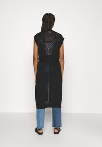 Cárdigan negro hasta la rodilla con una parte trasera semi-translúcida de estampado y mangas cortas, combinado con jeans de mezclilla anchos y deshilachados y sandalias.