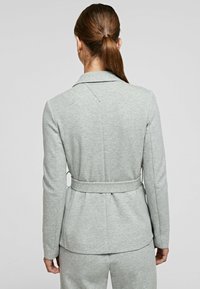 šedá, přiléhavá bunda s límcem, páskem v pase a jemnou strukturou. Obsahuje rozparek vzadu a čistý, minimalistický design.