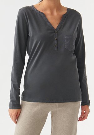 Kobieta ubrana w ciemnoszarą, długorękawową koszulę z dekoltem w serek, zapinaną na guziki z kieszonką na piersi, w połączeniu z beżowymi spodniami.