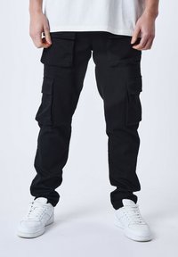 Pantalones cargo negros con varios bolsillos laterales, corte ajustado y tejido resistente. Combinados con zapatillas blancas sobre un fondo neutro.