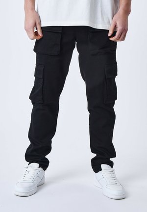 Pantalones cargo negros con varios bolsillos laterales, corte ajustado y tejido resistente. Combinados con zapatillas blancas sobre un fondo neutro.