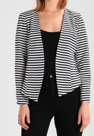 Kvinde iført sort-hvid stribet åben blazer over en sort top og sorte bukser, stående mod en ensfarvet baggrund.
