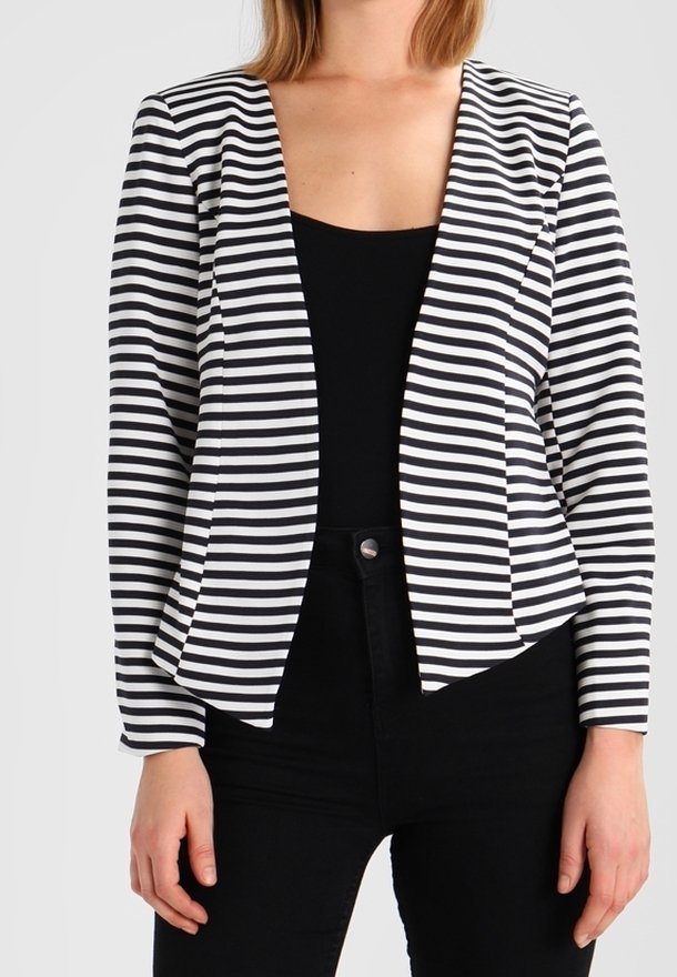 Femme portant un blazer ouvert à rayures horizontales noires et blanches sur un haut noir et un pantalon noir, debout devant un fond uni.