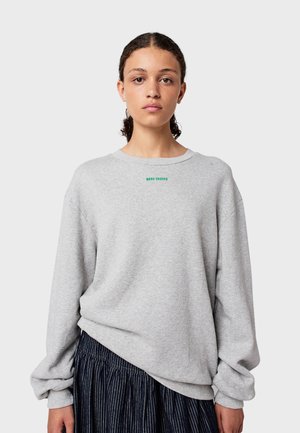 Felpa grigia con scollo rotondo, spalle cadenti e polsini a coste. Presenta del testo verde piccolo sul petto. Indossata con una gonna plissettata blu navy.