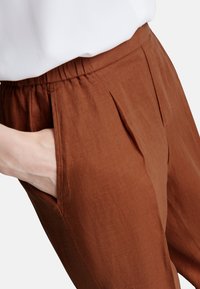 Hnědé lněné kalhoty mají volný střih, elastický pas a boční kapsy se subtilními sklady, čímž ukazují strukturovaný povrch látky.