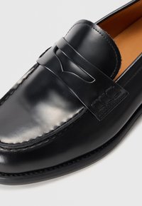 Mustadast nahast loafers, millel on õmmeldud mokassiini nina, dekoratiivne riba ja sile viimistlus koos pruuni sisepinnaga.