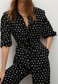 Schwarzer Jumpsuit mit weißen Punkten, mit durchgehender Knopfleiste, hochgerollten Ärmeln, tailliertem Gürtel und seitlichen Taschen. Weicher Stoff.