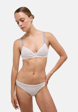 Femme portant un soutien-gorge blanc à motif en maille et une culotte assortie avec une bordure bleu clair, debout avec une main sur la hanche, regardant vers le bas.