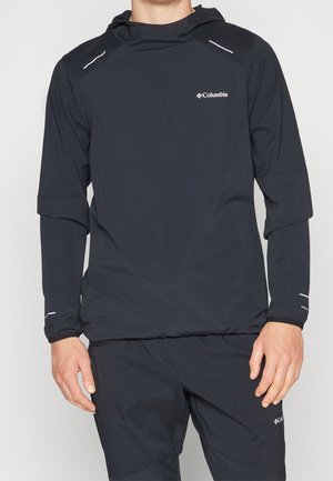 Homme portant une veste à capuche noire Columbia et un pantalon assorti sur un fond uni, montrant le haut du corps et les mains.