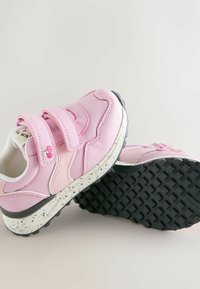 Rózsaszín sportcipők sima szintetikus felsőrésszel, dupla tépőzáras pántokkal és texturált fekete gumitalppal, fehér pöttyös középtalppal.