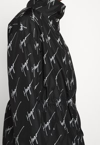 Czarna kurtka z białym napisem "Hugo" w stylu skryptu, z wysokim kołnierzem, elastycznym pasem i detalami z zatrzaskami widocznymi z boku.