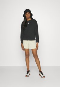 Sweatshirt noir avec logo, shorts beige clair, et chapeau seau noir. Les chaussures présentent un mélange d'accents gris, blancs et rouges.
