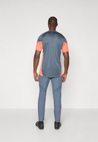 Grå atletisk t-shirt med orange detaljer, sammen med blå bukser. T-shirten har korte ærmer og en afslappet pasform, mens bukserne har tætsiddende ben.