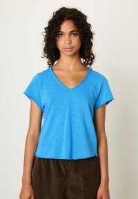 Modra bombažna majica s črnim vratnim izrezom, kratkimi rokavi in neurejenim robom, prikazana na modelu s curly lasmi.