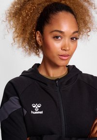 Svart zip-up hoodie med en mjuk textur, som har en grå detalj på ärmarna och en vit logotyp på bröstet.