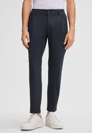 Man die marineblauwe, op maat gemaakte broek en witte sneakers draagt, staand tegen een effen lichte achtergrond.