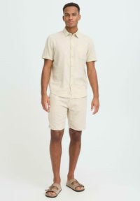 Chemise beige en lin à manches courtes avec une poche poitrine, assortie à un short coordonné. Portez avec des sandales marron dotées d'une double bride.