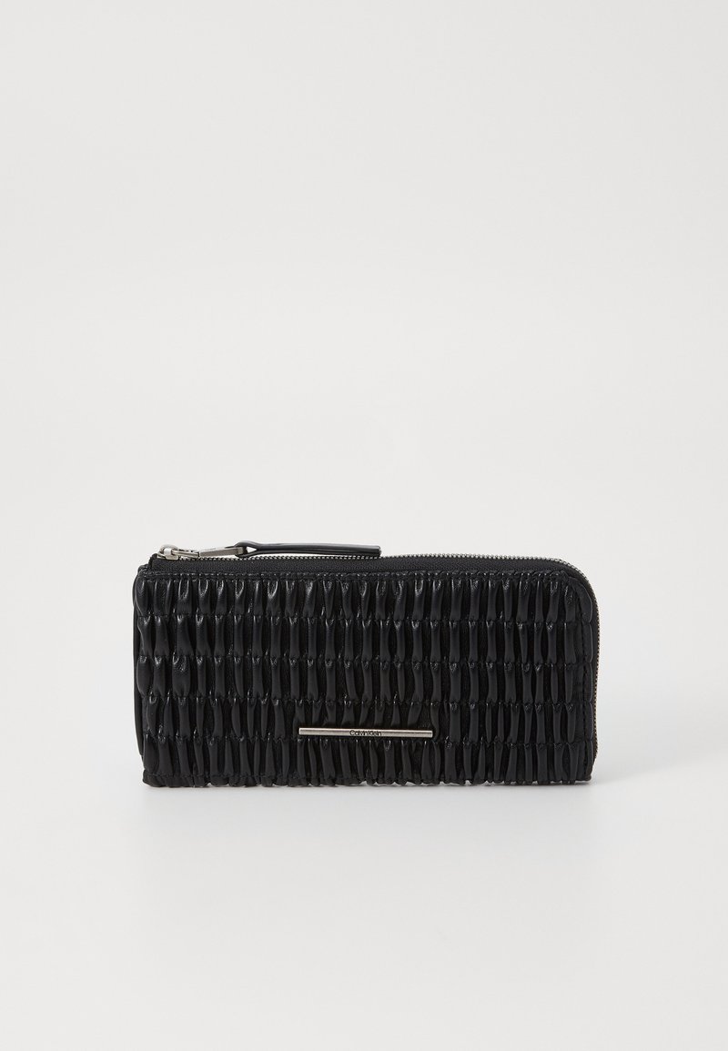Portefeuille noir avec une finition texturée, un design plissé, une fermeture éclair et une bande métallique à l'avant. Matériau lisse.