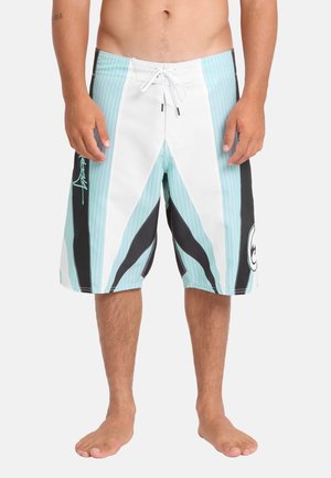 Homme portant un short de bain bleu clair, blanc et noir arrivant aux genoux avec un cordon, debout pieds nus sur fond blanc.