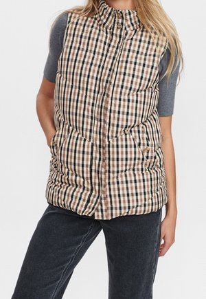 Femme portant une veste matelassée à carreaux beige, marron et noir par-dessus un haut gris à manches courtes et un pantalon sombre, debout avec une main dans la poche.