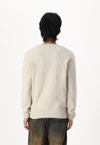 Personne aux cheveux foncés vue de dos, portant un pull tricoté crème à motif losange et un pantalon brun foncé délavé avec de grandes poches arrière.