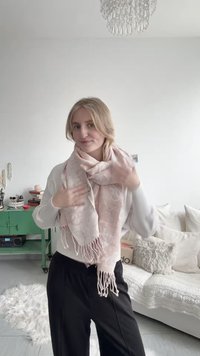 Blonde woman in white sweater and black pants adjusting a pink patterned scarf, standing in a living room with white couch and green cart.