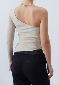 One-shoulder top in metallic beige fabric with ribbed texture, paired with black pants featuring back pockets and a smooth waistband.