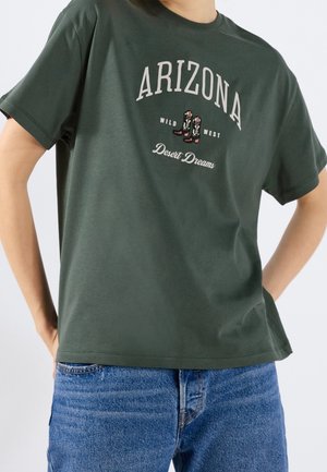 Mørkegrøn T-shirt med "ARIZONA", cowboy-støvle grafik, "Wild West" og "Desert Dreams" tekst, båret med blå denimjeans.