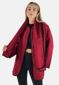 Abrigo rojo con ribetes negros, diseño oversize, tejido texturizado, cinco botones, frente abierto y mangas amplias, llevado sobre una blusa y pantalones negros.