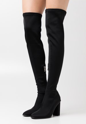 Bottes noires à talons hauts jusqu'aux genoux avec des fermetures éclair sur le côté portées sur des jambes nues devant un fond clair.