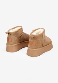 Botas de tobillo de ante beige con forro de piel de oveja blanca suave, suelas de goma planas y un pequeño tacón trasero. Presenta un acento de cuero de marca en la parte posterior.