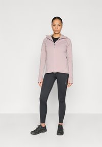 Casaco rosa claro com fecho de correr e capuz, usado sobre uma t-shirt preta e combinado com leggings pretas. Calçado preto com detalhes texturizados.