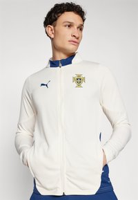Jackett bianco con accenti blu navy, caratterizzato da un logo Puma e un emblema del Portogallo sul petto. Realizzato in tessuto performante testurizzato.