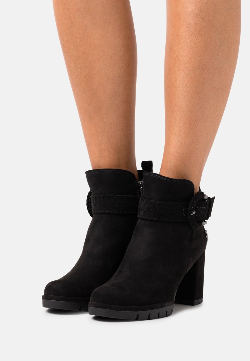 Bottes courtes en daim noir avec talons épais, lanière tressée et détail de boucle portées sur des jambes nues sur fond blanc.